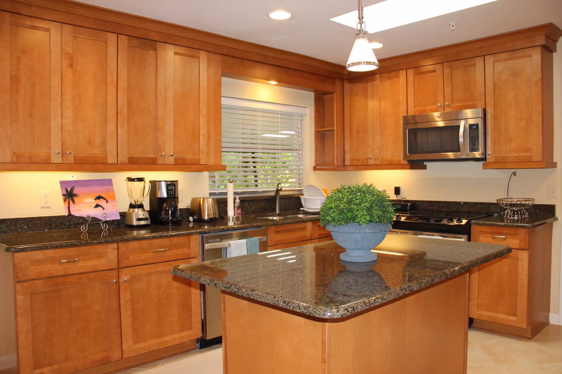 A modern kitchen with wooden cabinets, granite countertops, and a central island. The kitchen features a stainless steel microwave, stove, toaster, blender, coffee maker, and a small potted plant on the island. A window with white blinds is above the sink.