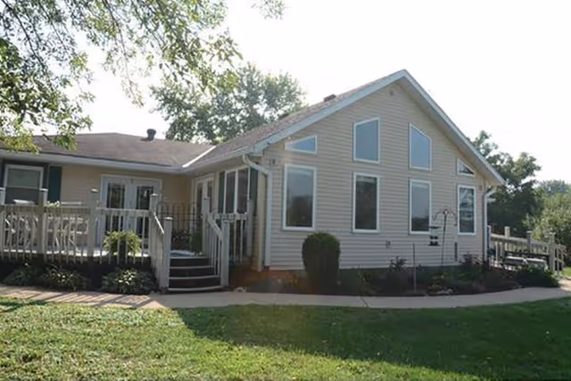 Front exterior of a beige single-story house with large windows, a raised deck/porch, and a grassy lawn.