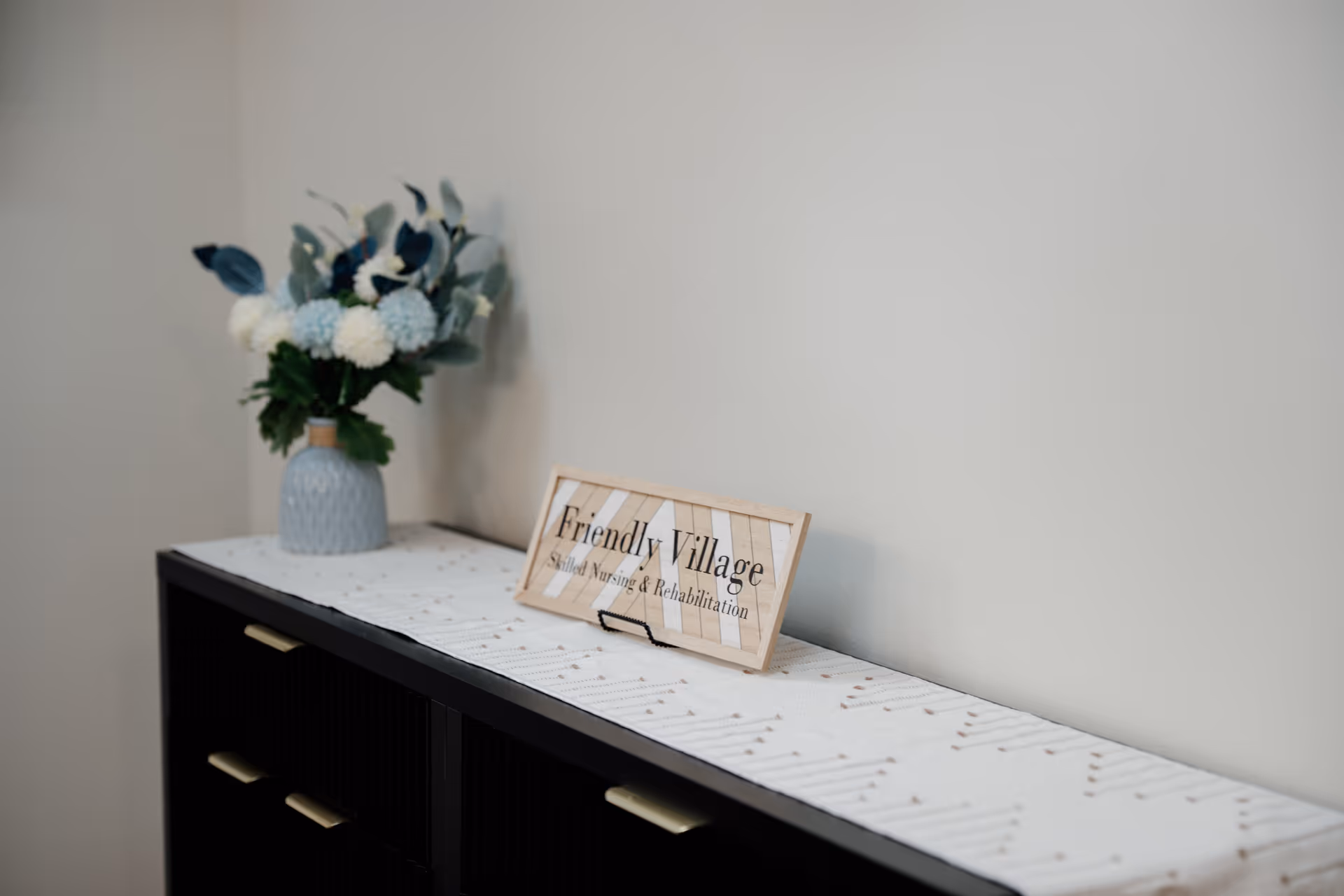 A vase of flowers sits on a console table beside a 'Friendly Village' sign.