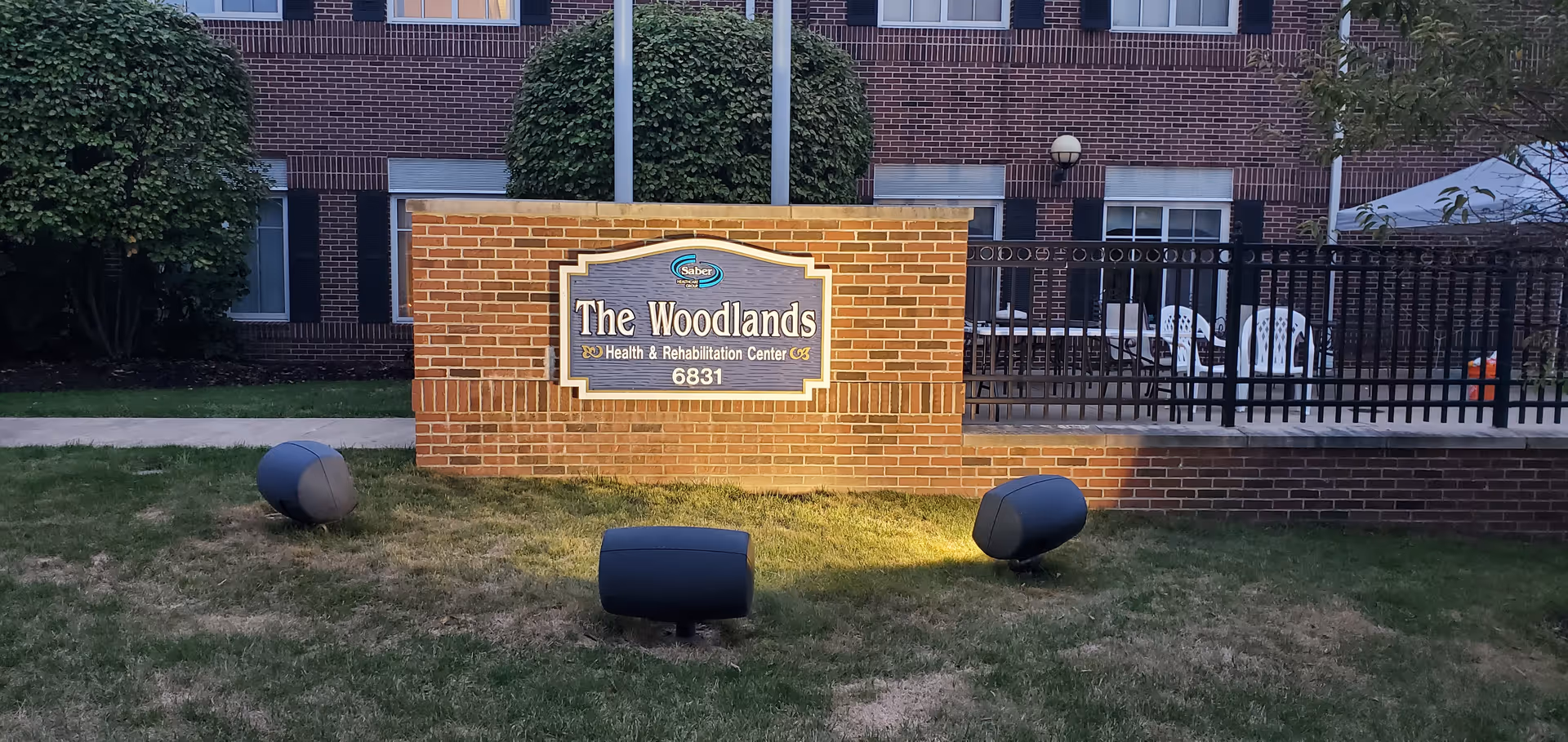 Brick sign reading The Woodlands Health & Rehabilitation Center with lawn and building.