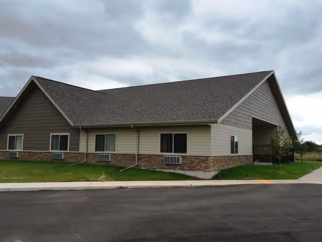 Exterior view of a single-story building with a gable roof, beige and brown siding, several windows with air conditioning units below them, and a paved driveway in front under a cloudy sky.