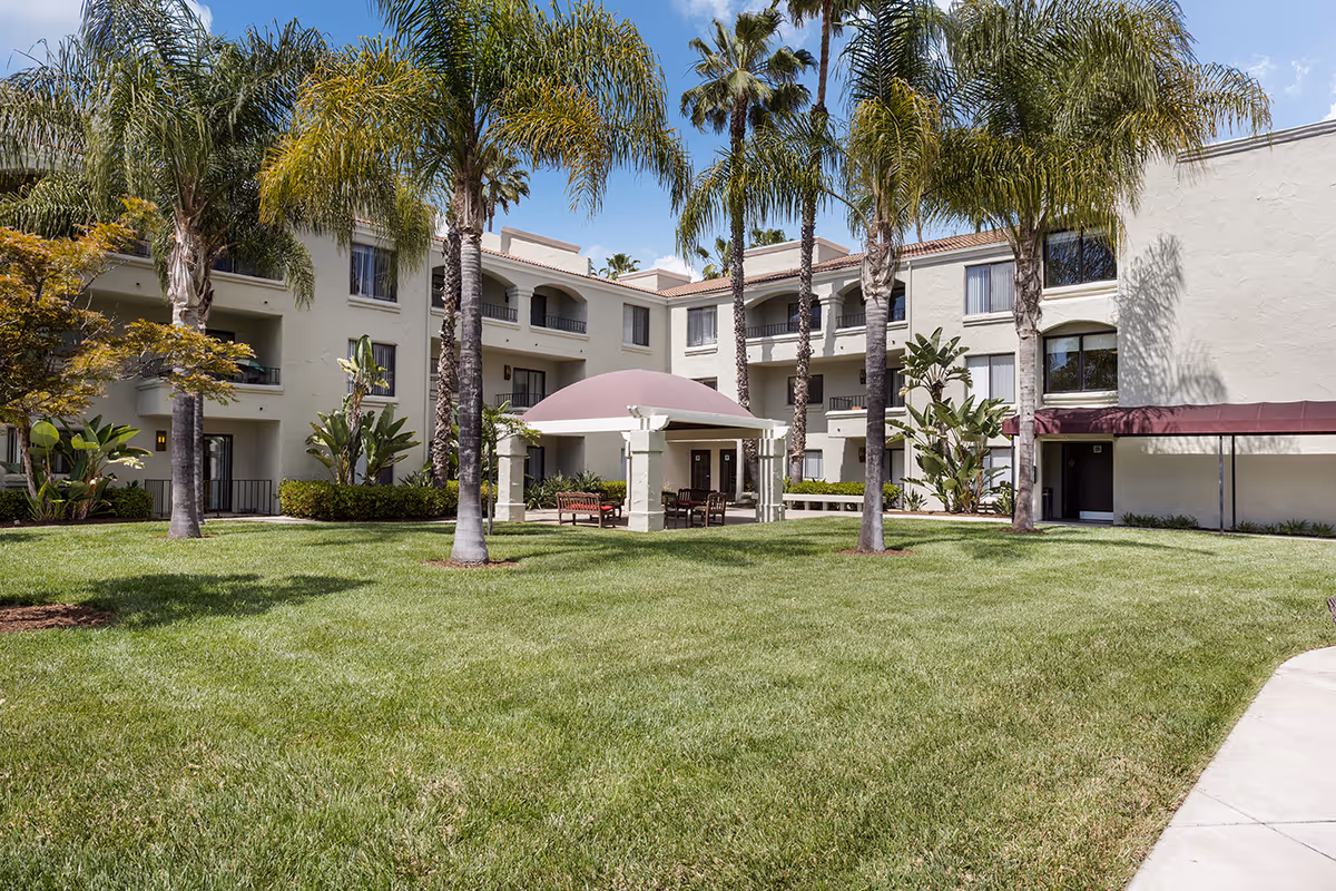 Green lawn and palm trees in a courtyard in front of a three-story beige senior living building with a covered seating area.