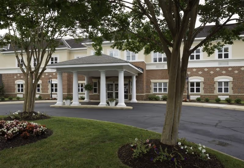 Front entrance of a brick senior living building with a covered porte-cochère supported by white columns, landscaped beds, and trees.