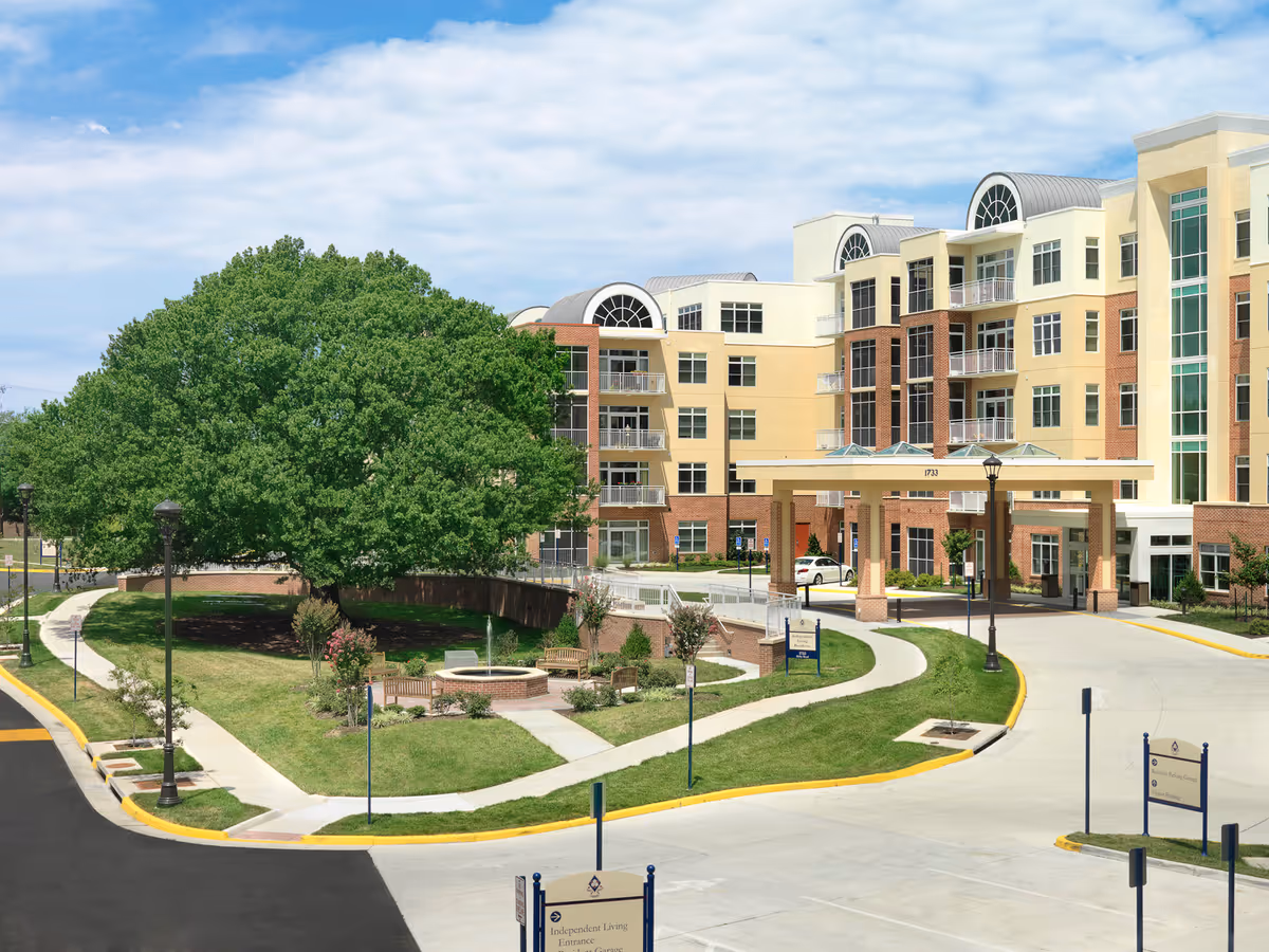 Exterior view of the Arleigh Burke Pavilion at Vinson Hall Retirement Community showing a multi-story building with a covered entrance, large windows, and balconies. In front of the building is a landscaped area with a large tree, benches, a circular brick planter, and walking paths. The sky is partly cloudy.