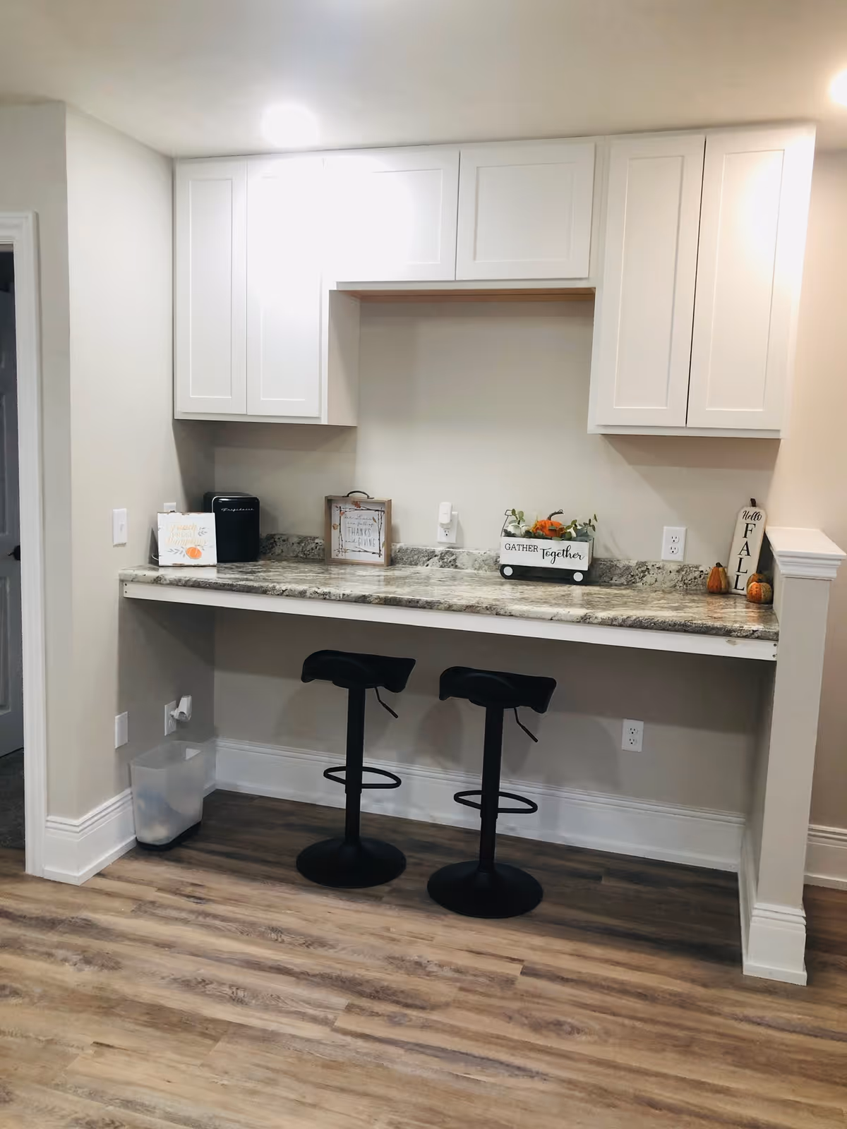 A small kitchen or dining nook area with a granite countertop bar, two black bar stools, white upper cabinets, and fall-themed decorations including small pumpkins and signs that say 'Gather Together' and 'Hello Fall'. The floor is wood, and there is a trash bin under the counter on the left side.