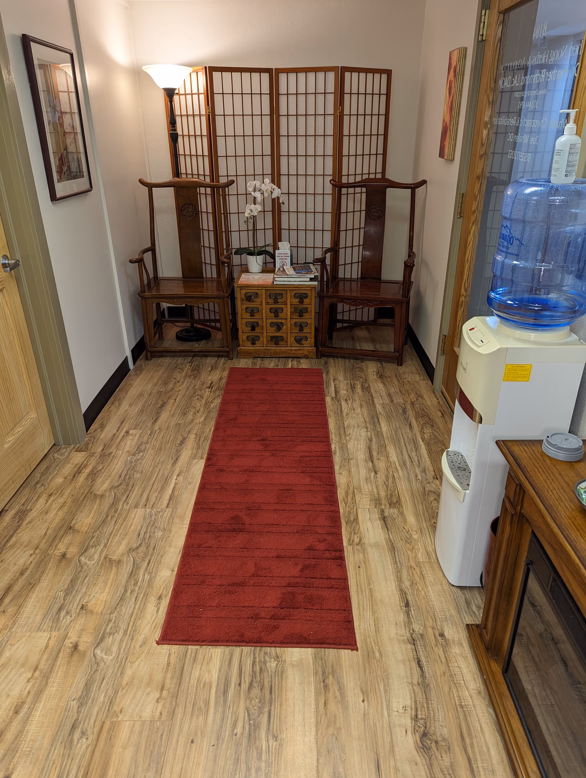Wooden chairs flank a cabinet and folding screens in a hallway, with a red runner rug.