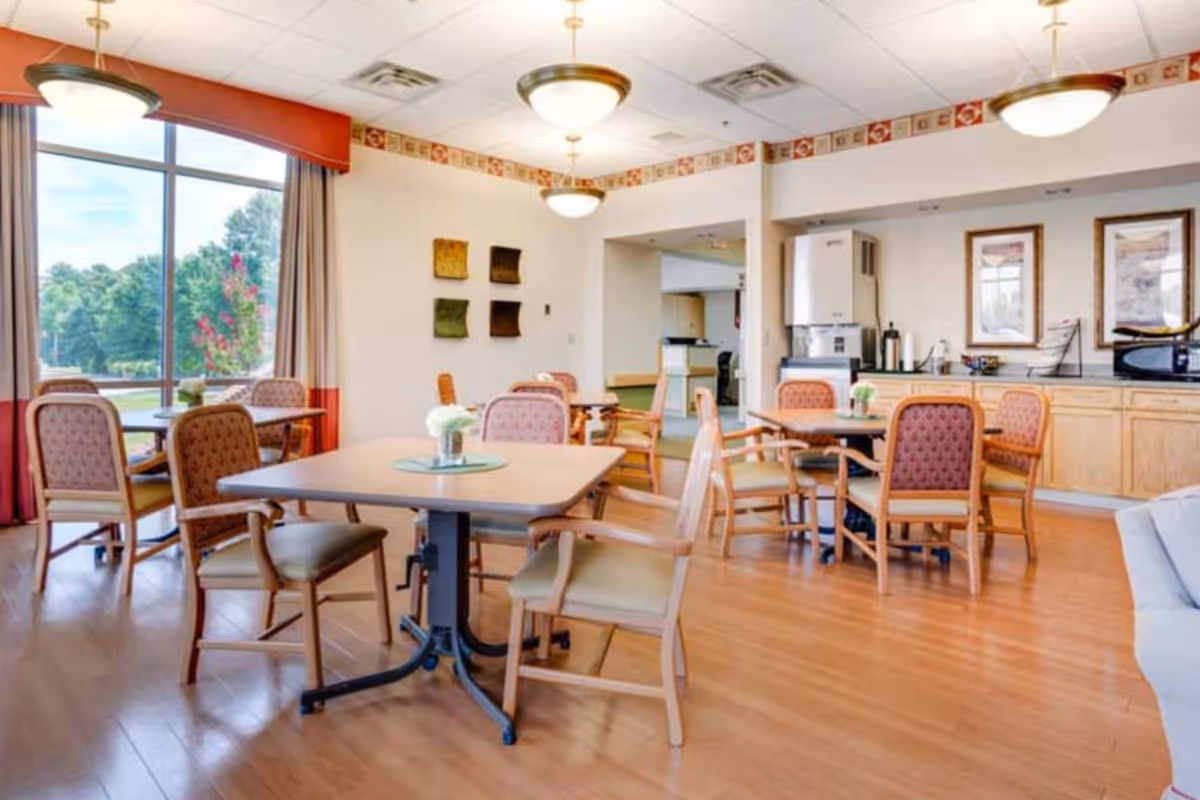 Bright dining room with multiple tables and chairs, large windows, and a kitchenette area in the background.