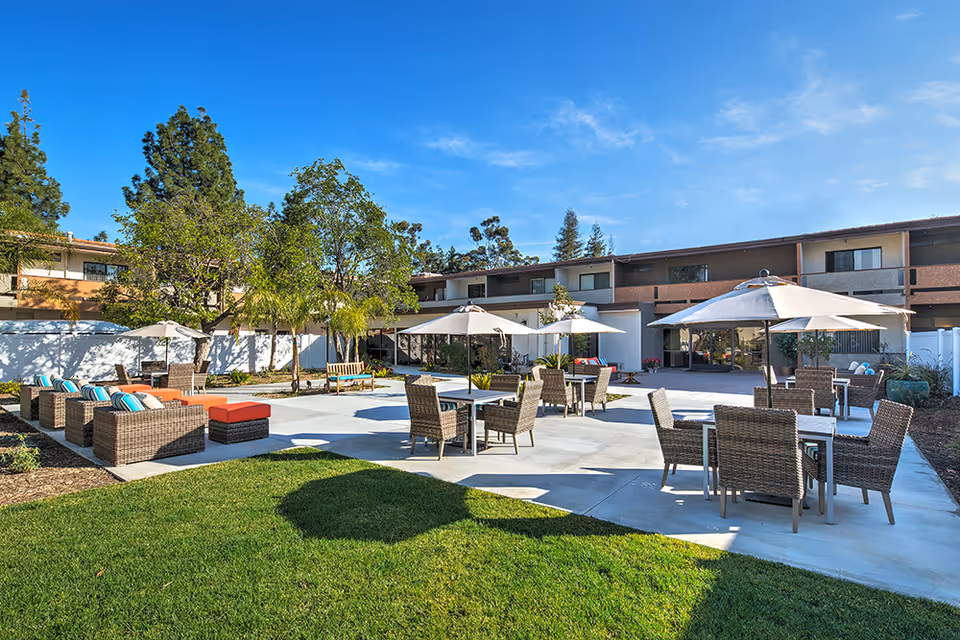 Outdoor patio area at Canyon Trails at Topanga Senior Living with multiple wicker chairs and tables under large white umbrellas, surrounded by green grass, trees, and a two-story building in the background under a clear blue sky.