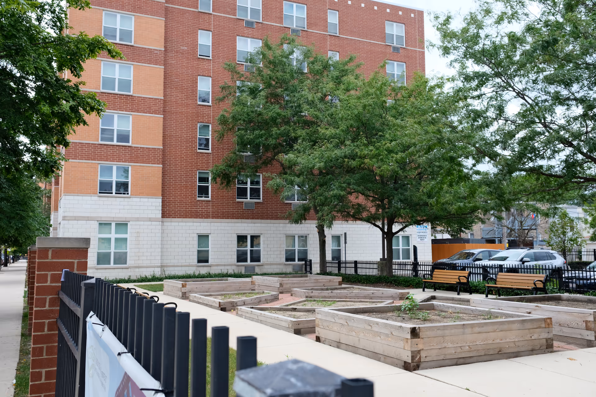Brick multi-story building with raised wooden garden beds, benches, and trees in a fenced front courtyard.