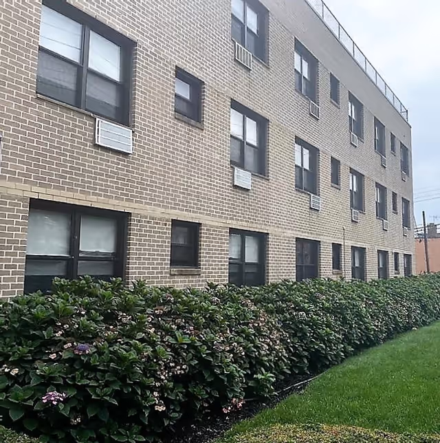 Exterior view of a multi-story brick building with several windows, air conditioning units installed below some windows, and a neatly trimmed hedge with small flowers in front of the building. The sky is overcast.