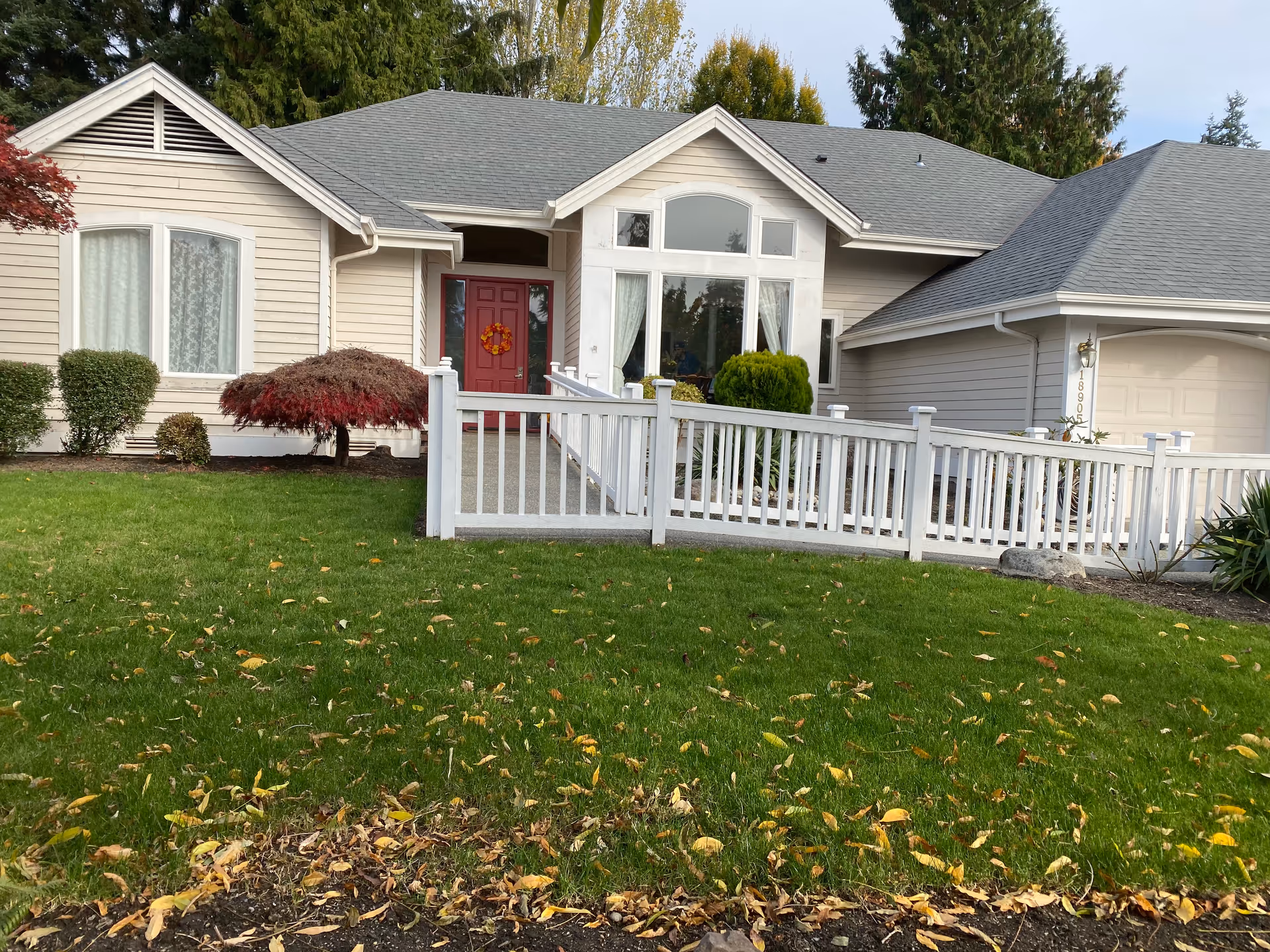 A single-story beige house with white railing, red front door, and green lawn.