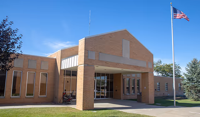 Brick hospital-style building with glass entrance, porte-cochere, and a flagpole under a blue sky.
