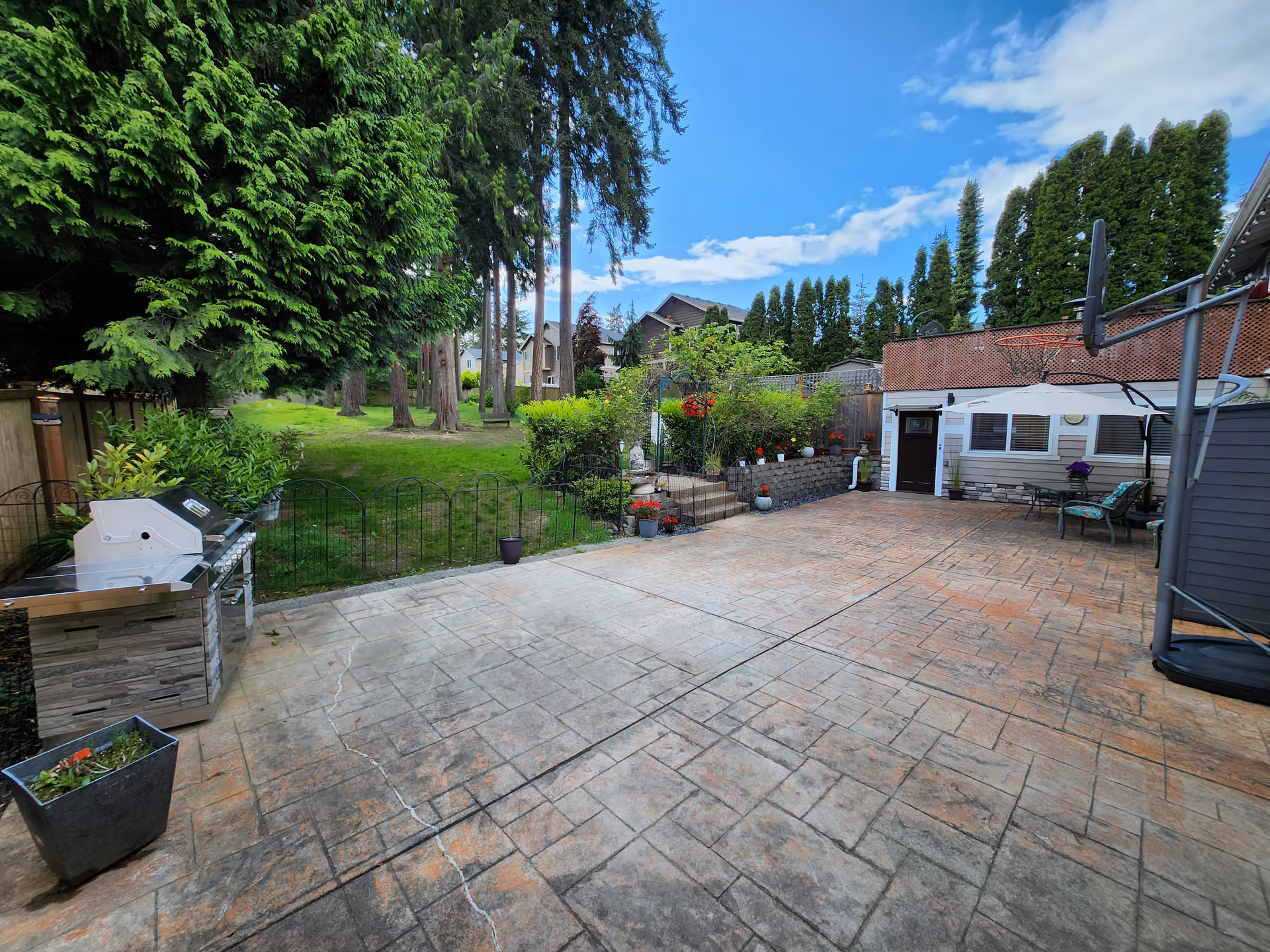 Spacious paved backyard patio with grill, plants, and seating area beside a small building.