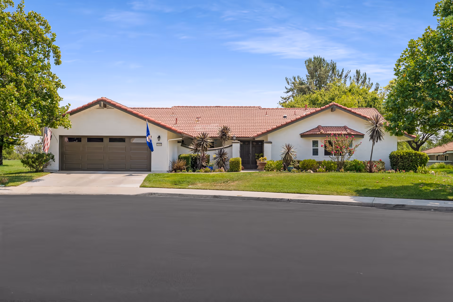 Single-story stucco building with a red tiled roof, two-car garage, front lawn and trees under a blue sky.