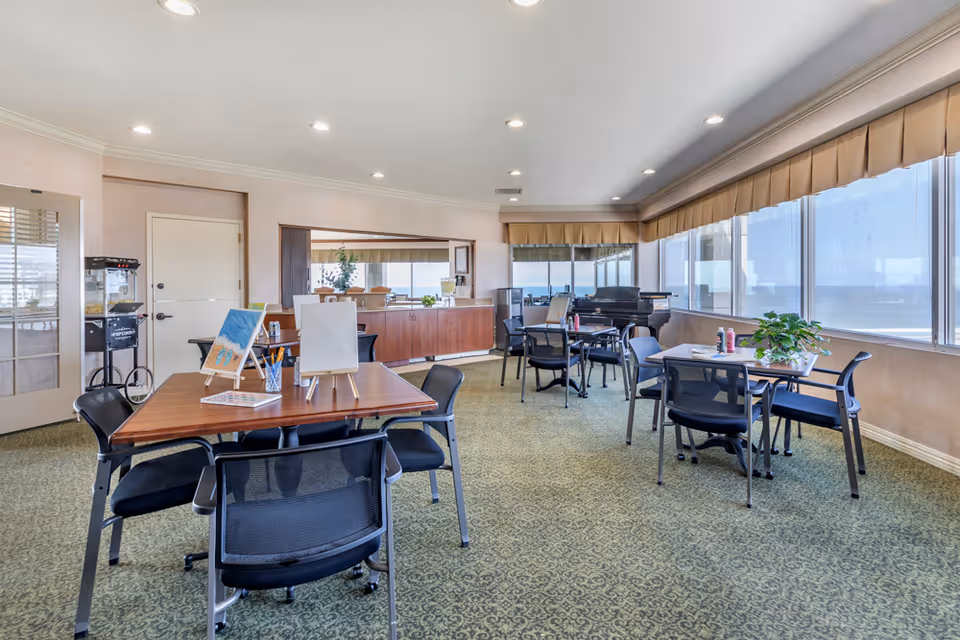 Interior common area with tables, chairs, a piano, and large windows in a senior facility.
