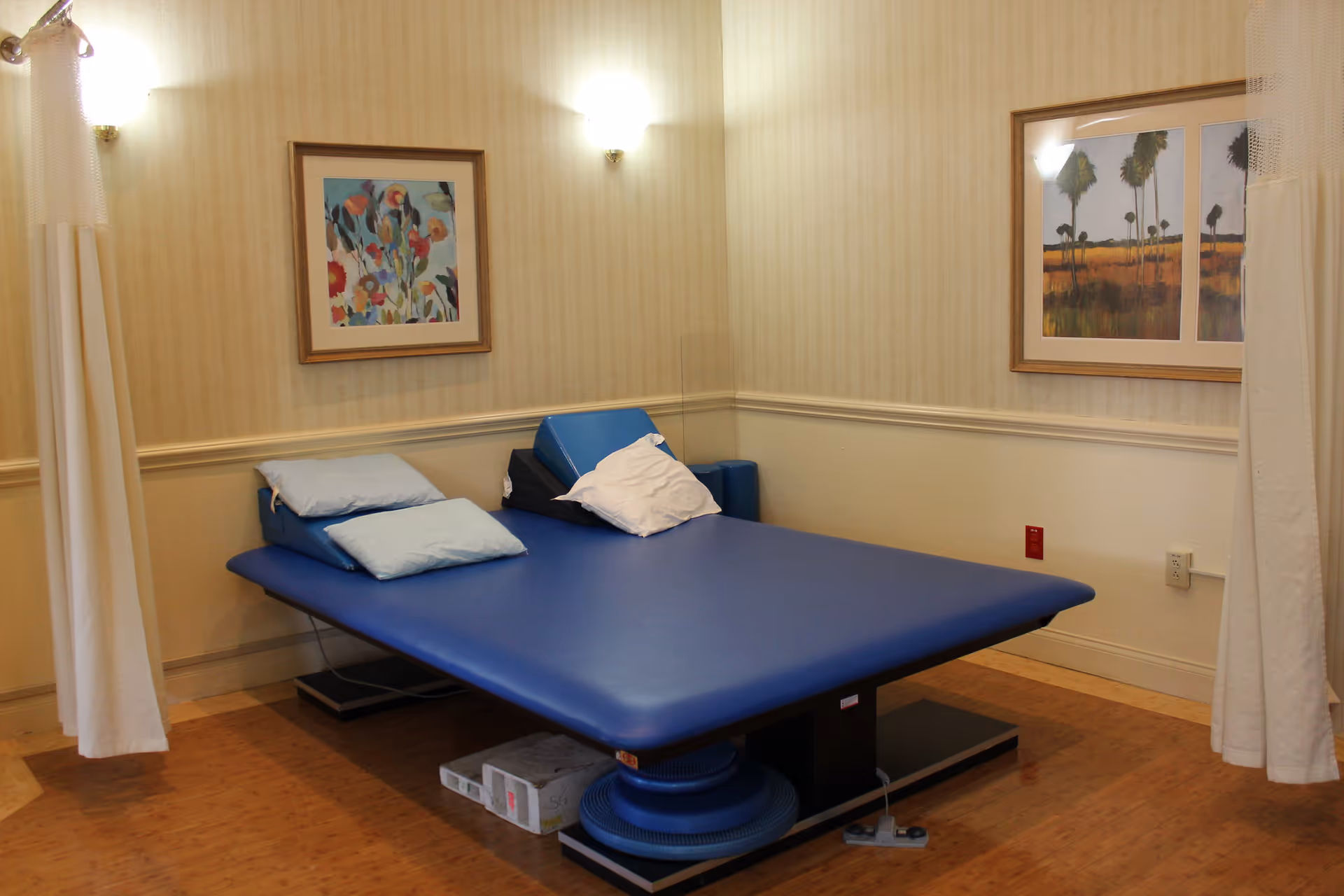 A therapy or treatment room with a large blue padded table, several pillows, and two framed paintings on the walls. The room has light-colored wallpaper, wooden flooring, and white curtains partially drawn on either side.