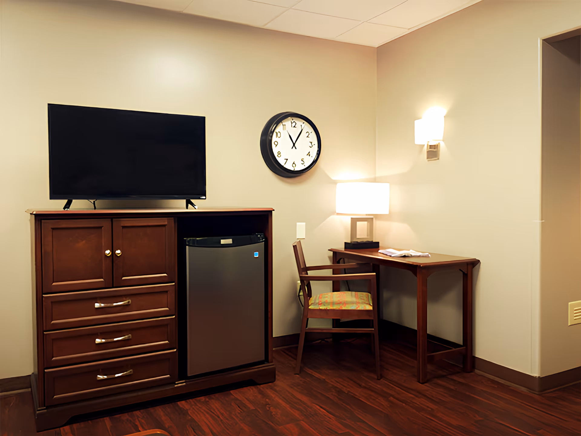 Small sitting area with a TV on a wooden cabinet, a mini fridge, a wall clock, and a desk with chair and lamp.