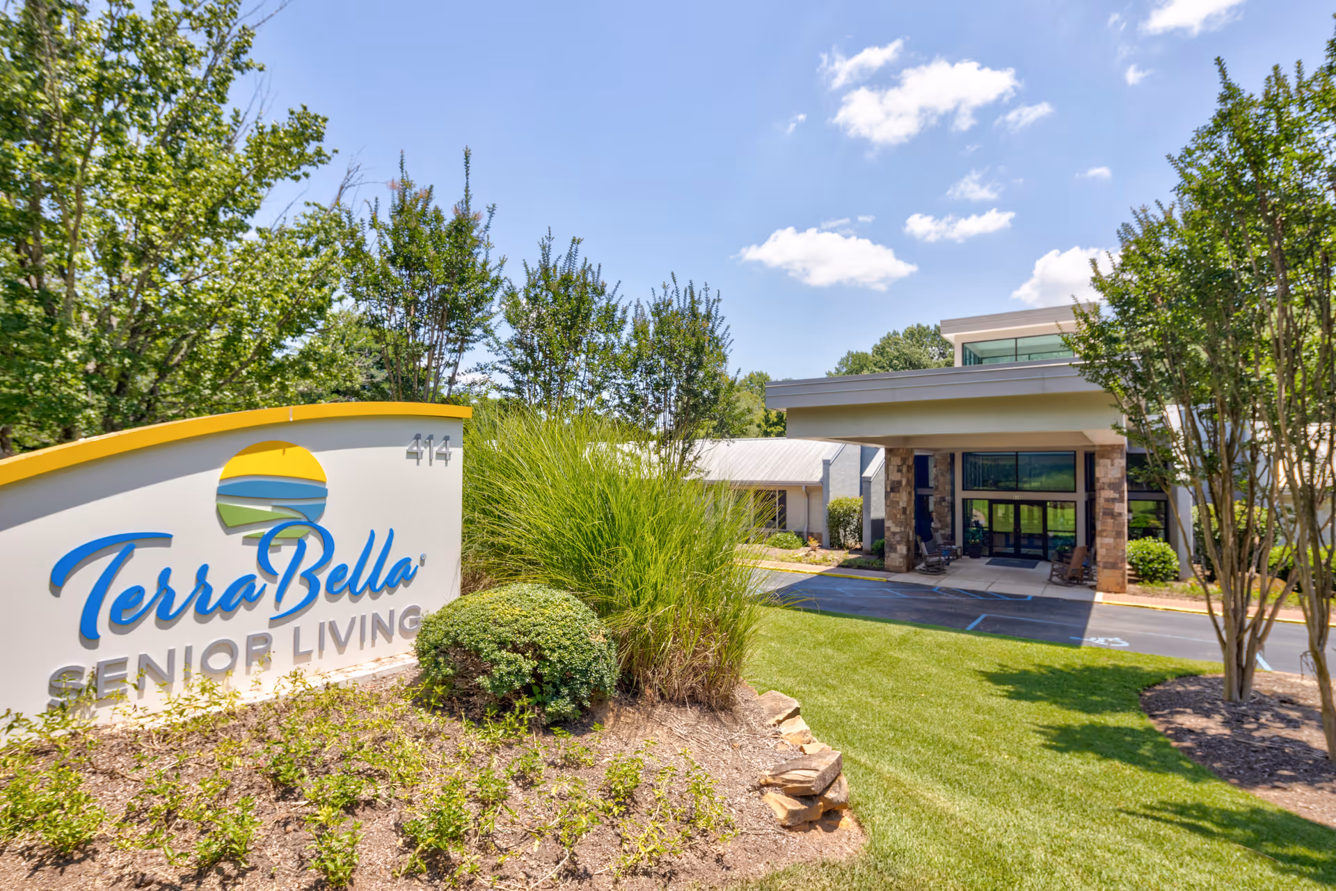 Exterior view of TerraBella Senior Living facility showing the entrance with a covered driveway, surrounded by green trees and landscaping under a blue sky with some clouds. A large sign with the TerraBella logo and the words 'Senior Living' is prominently displayed in the foreground.