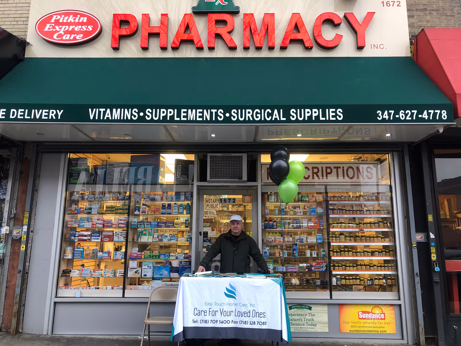 A man stands behind a table outside a storefront pharmacy with balloons.