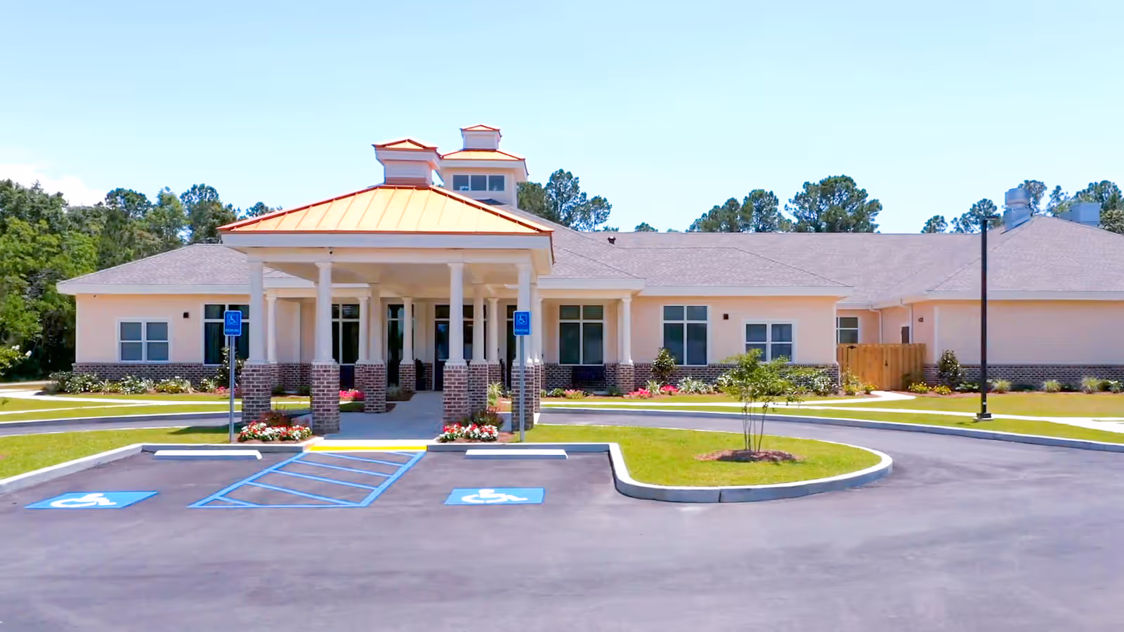 Front exterior view of The Homestead Assisted Living Community building with a covered entrance supported by white columns and brick bases. The building has a light-colored facade with multiple windows and a gray roof. There are two handicapped parking spaces in front, surrounded by landscaped grass and small plants, with trees in the background under a clear blue sky.
