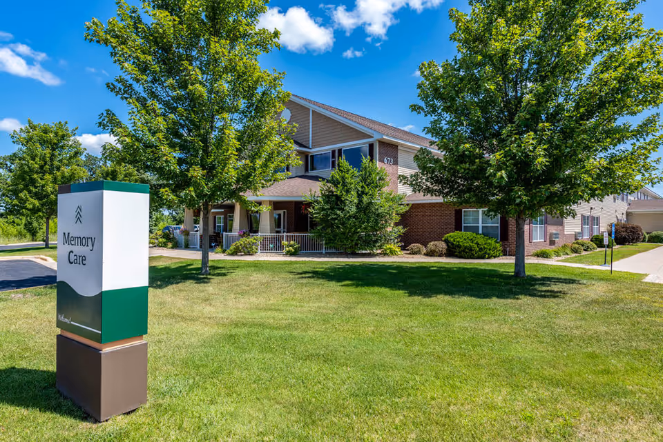 Exterior view of a senior living facility building with a green lawn and trees under a blue sky with some clouds. A sign in the foreground reads 'Memory Care'.