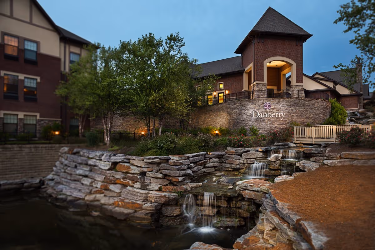 Exterior view of Danberry at Inverness facility during dusk, showing a multi-story building with lit windows, a stone retaining wall, landscaped greenery, and a small cascading waterfall feature in the foreground.