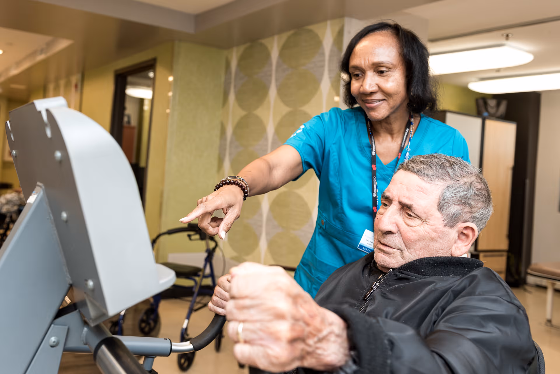 A healthcare worker in teal scrubs assists an elderly man using an exercise machine in a rehabilitation center. The man is seated and gripping the machine's handles while the worker points at the machine, providing guidance. The background shows a walker and a room with patterned wallpaper and furniture.