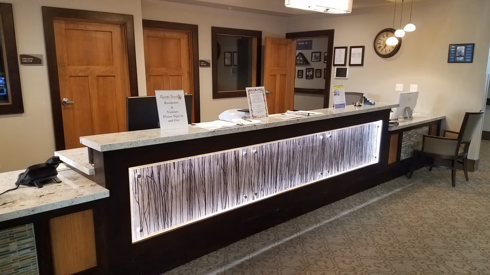 Reception desk in a senior living lobby with illuminated panel and wooden doors behind.
