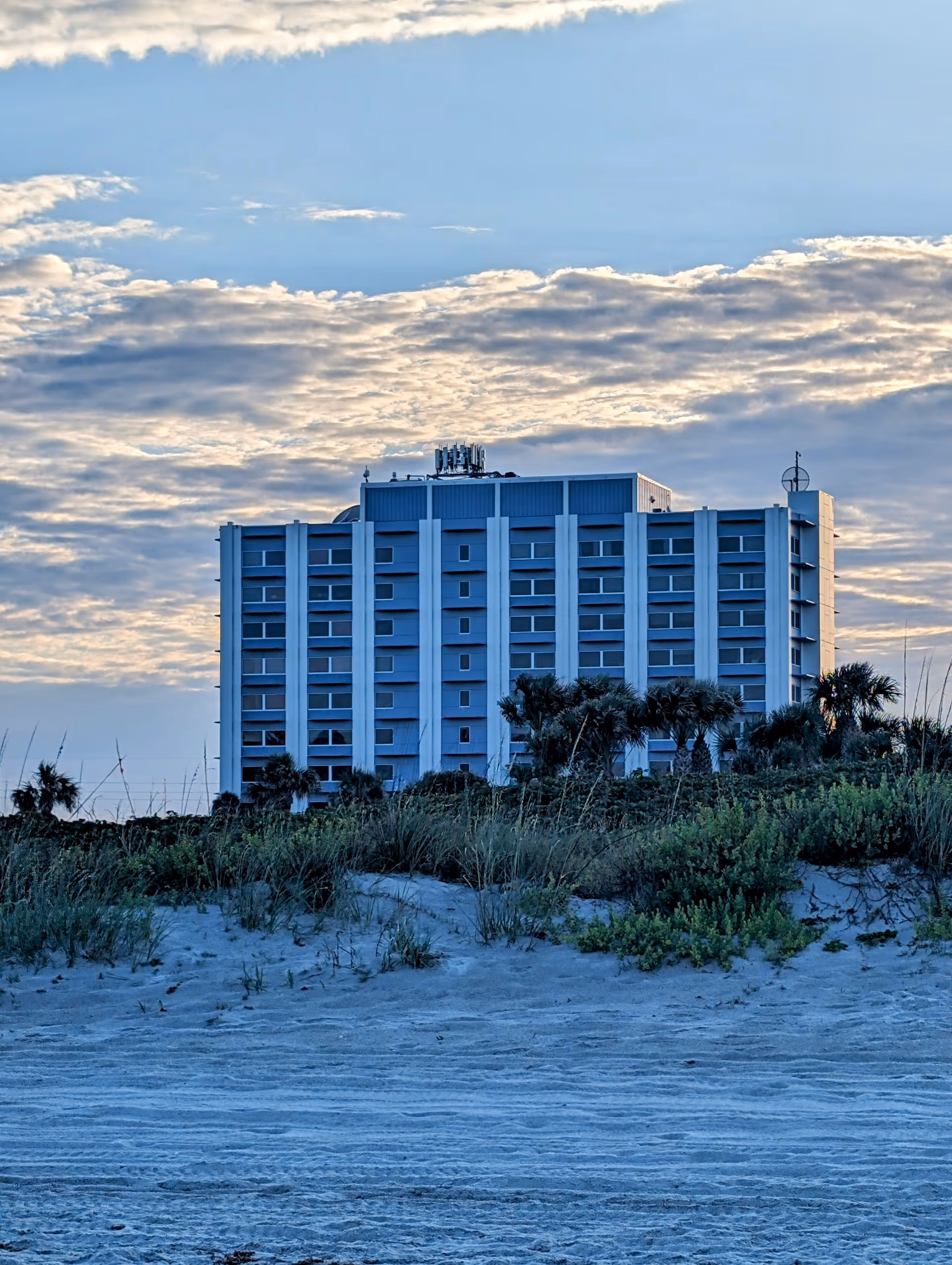 A tall white, multi-story building sits along a sandy beach with dunes.