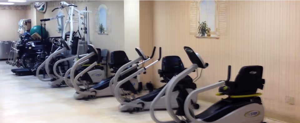 Indoor gym room with multiple exercise machines and seating against a light-colored wall.