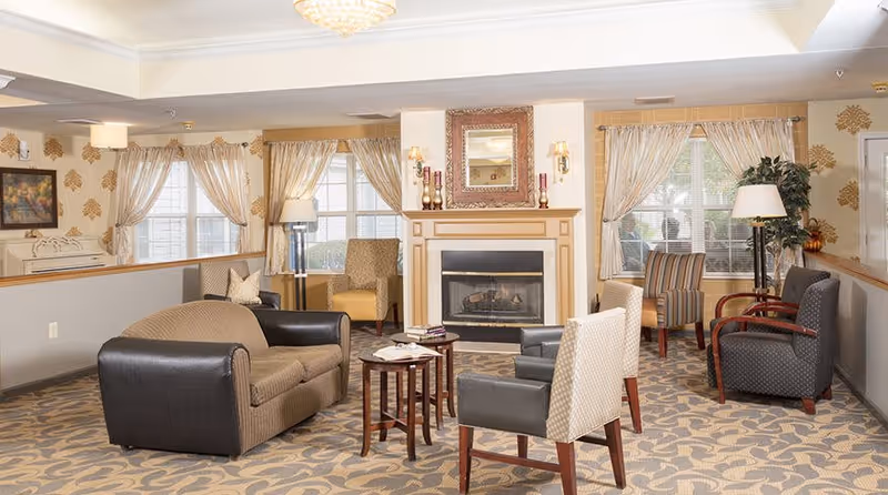 A cozy assisted living facility common area with multiple armchairs and a loveseat arranged around a fireplace. The room features patterned carpet, large windows with curtains, wall sconces, a framed mirror above the fireplace, and decorative lamps and plants.