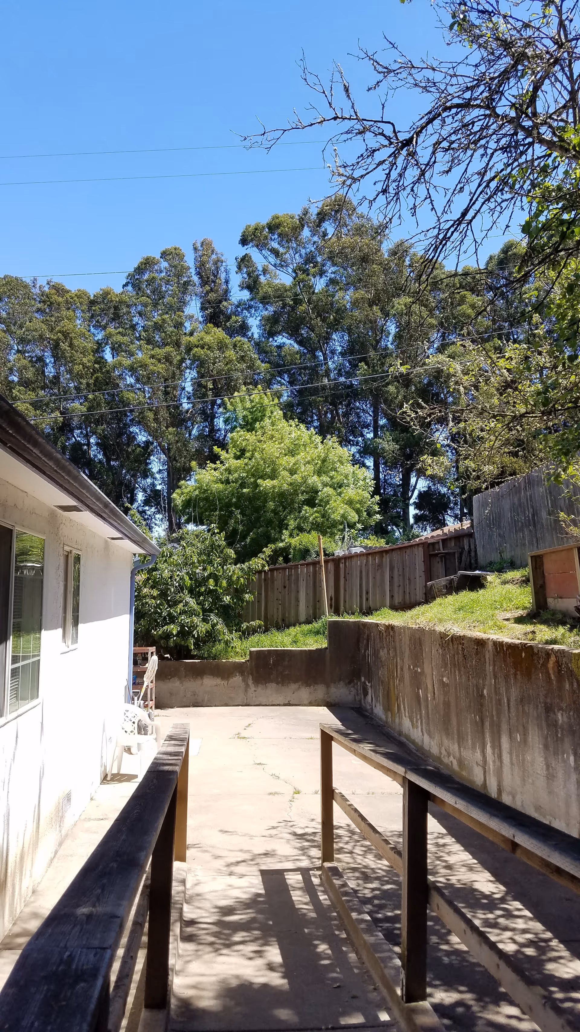 Outdoor concrete ramp and patio with white wall, railings, trees, and a wooden fence.
