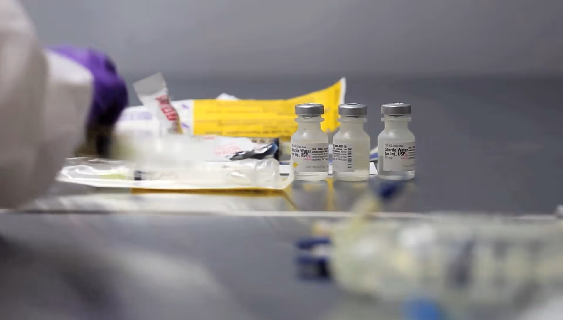 Medical vials and supplies on a blurred stainless steel table in a clinical interior.