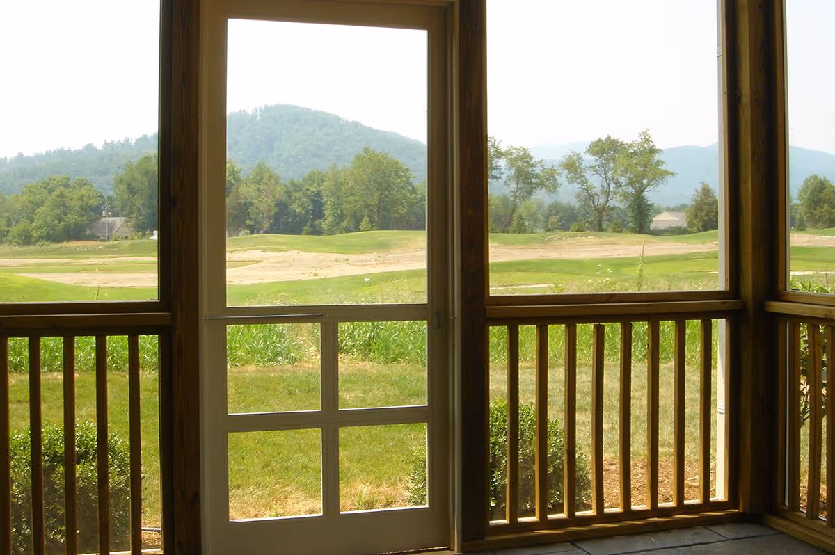 View through a screened wooden porch with a central screen door and railing onto a grassy lawn and distant tree-covered hills.