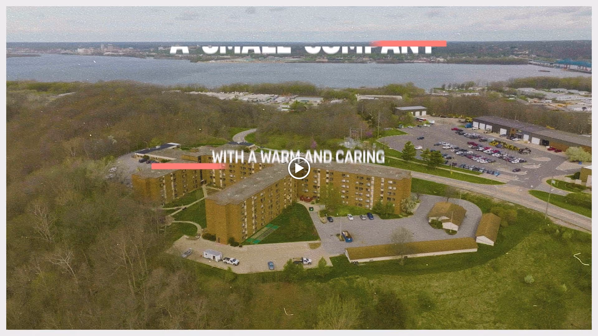 Aerial view of a large multi-story residential building surrounded by trees and parking lots, with a river and bridge visible in the background under a cloudy sky.