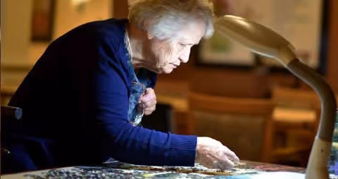 An elderly woman with white hair wearing a dark blue sweater is focused on assembling a jigsaw puzzle on a table under a bright lamp in a cozy indoor setting.