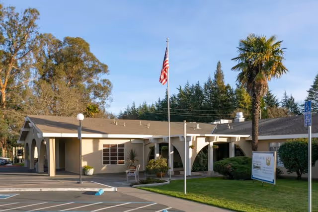 Front entrance of a single-story senior living facility with an American flag, palm tree, and parking spaces.