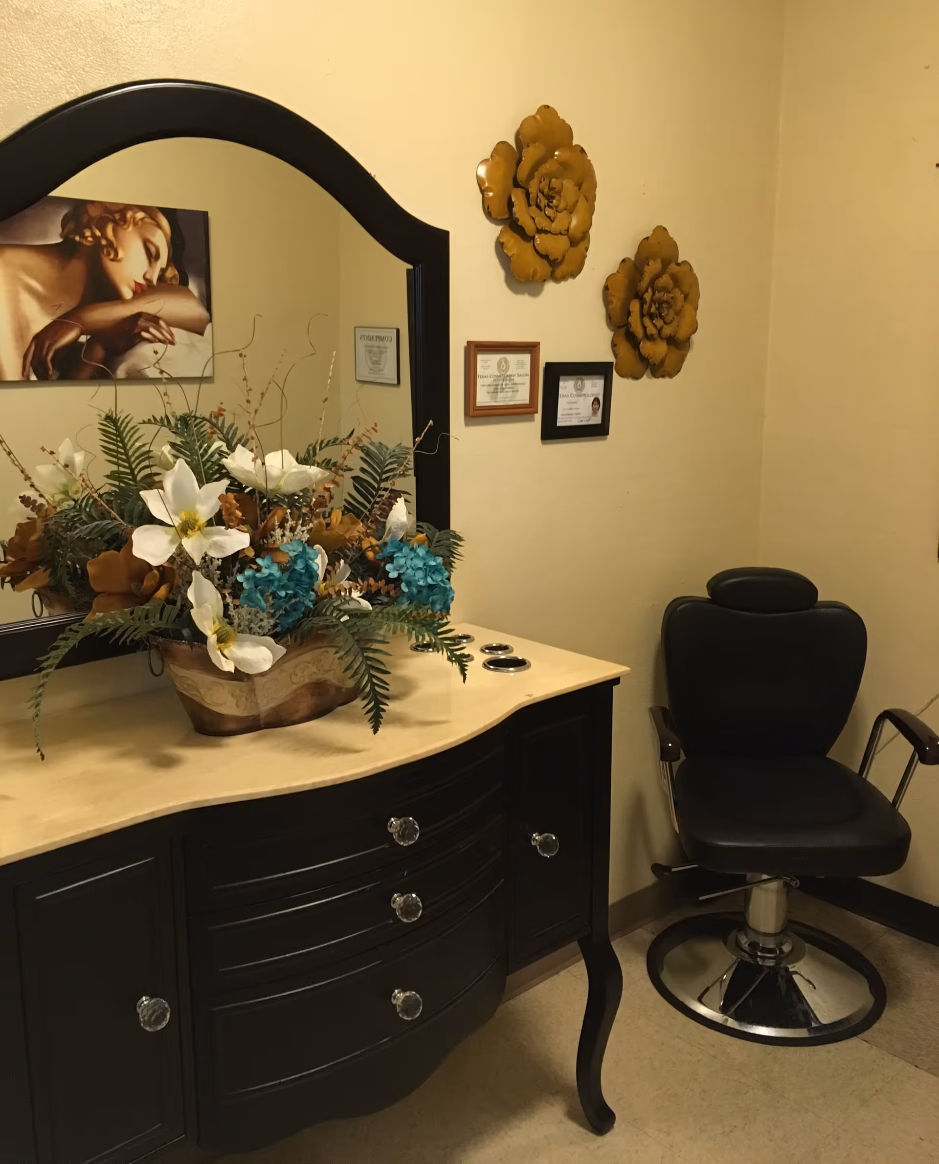 A small room with a black salon chair and a black dresser with a beige countertop. On the countertop is a decorative floral arrangement with white and blue flowers and green leaves. Above the dresser is a large mirror with a black frame. The wall has two large yellow flower decorations and three framed certificates or documents. A painting of a woman resting her head on her arm is partially visible in the mirror reflection.