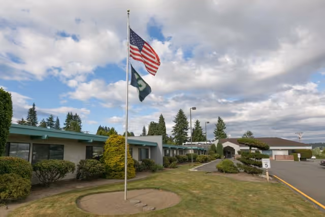 Low-rise care facility building with an American flag and a second flag on a flagpole, trimmed shrubs, and a driveway under a partly cloudy sky.