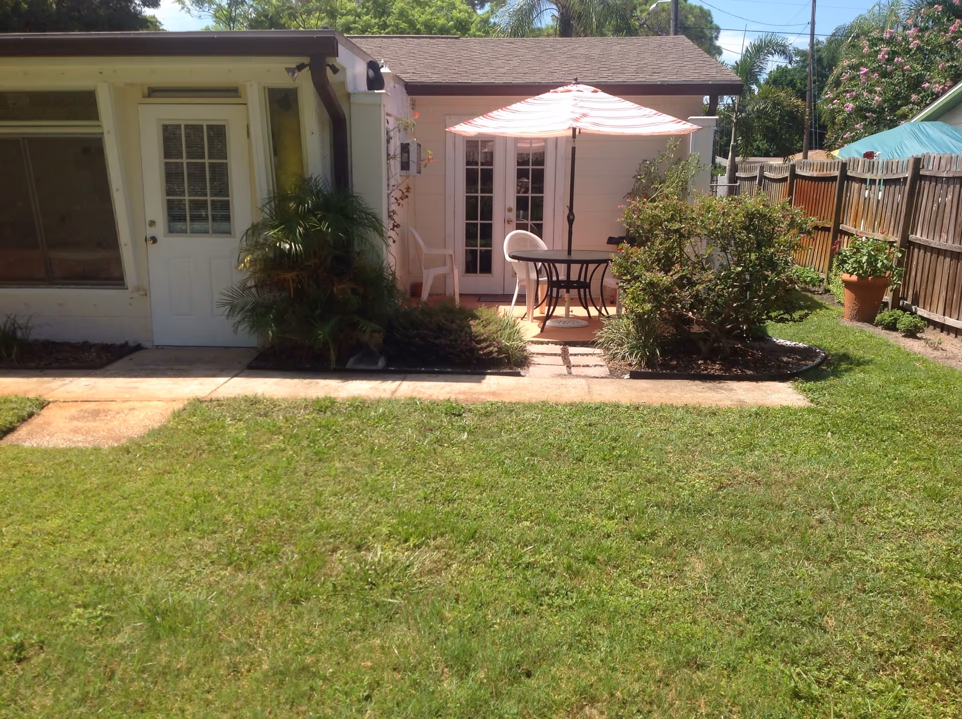 A backyard garden area with a small patio featuring a round table, two white chairs, and a large striped umbrella. The patio is adjacent to a white building with a door and windows. There are plants and bushes along the edges of the patio and a wooden fence on the right side.