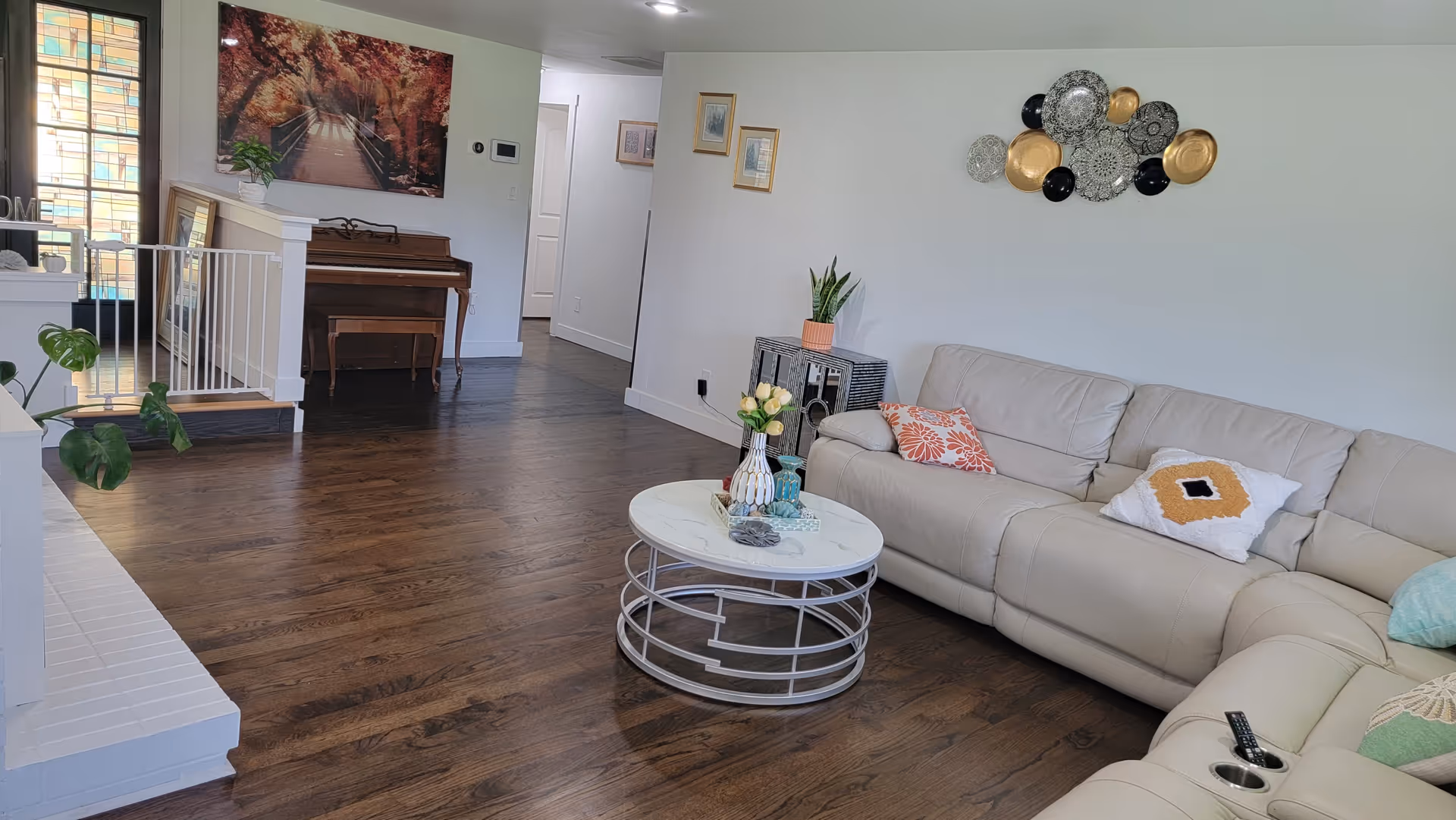 Bright living room with a beige sectional sofa, round coffee table, piano in the background, and dark hardwood floors.