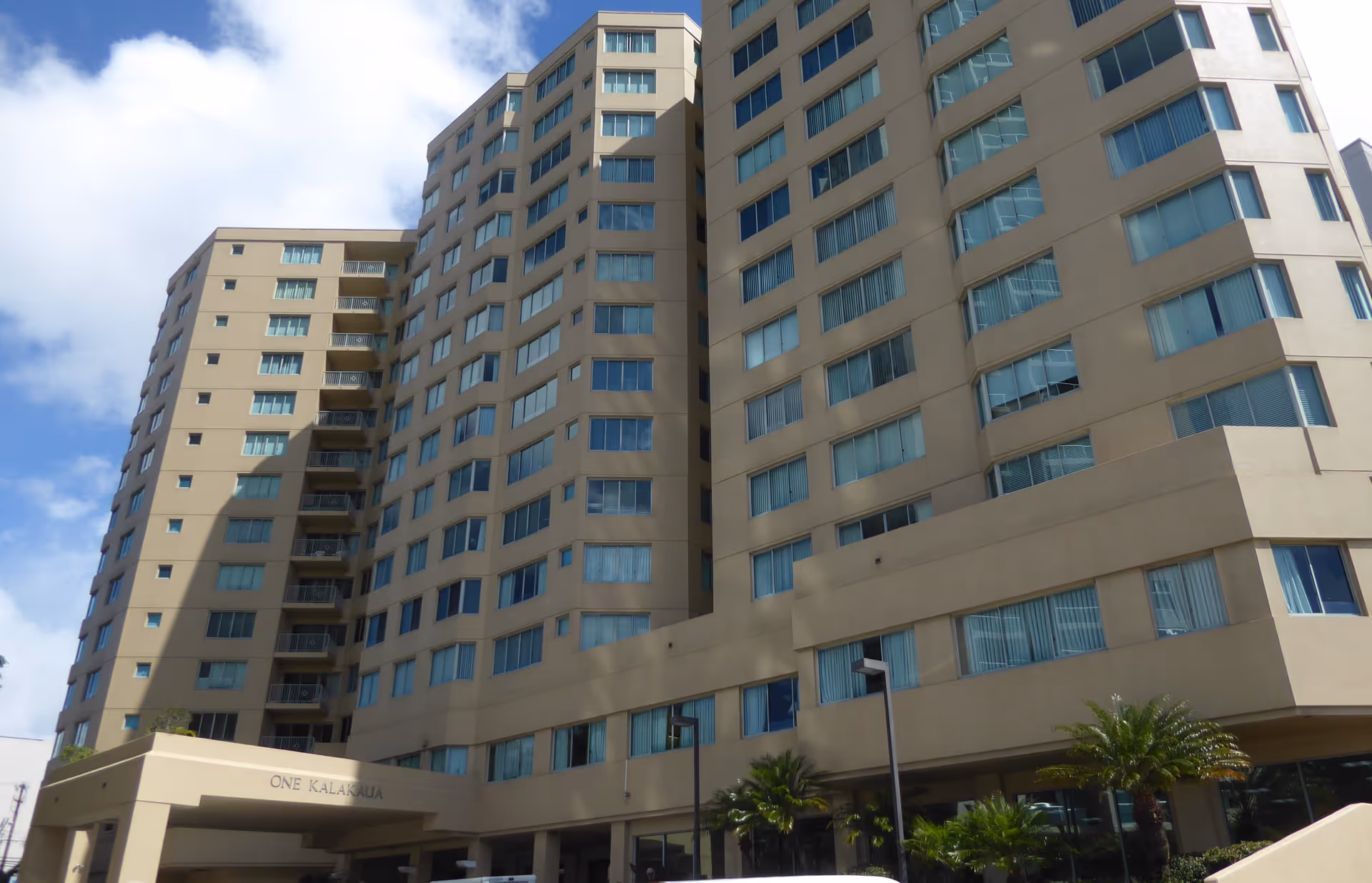 Tall beige Hale Ola Kino senior living building with many windows, palms, and covered entrance.