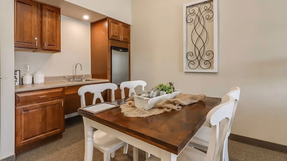 A small dining area with a wooden table and four white chairs. The table has a decorative centerpiece with a basket, greenery, and some dishes. In the background, there is a kitchenette with wooden cabinets, a sink, and a small refrigerator. A decorative wall hanging with a wrought iron design is mounted on the beige wall.