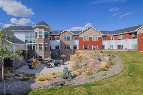 Exterior view of a senior living facility named The Homestead at Rochester, featuring a multi-story building with a mix of stone and red siding. The building surrounds a landscaped courtyard with patio tables and umbrellas, small trees, and shrubs under a blue sky with some clouds.