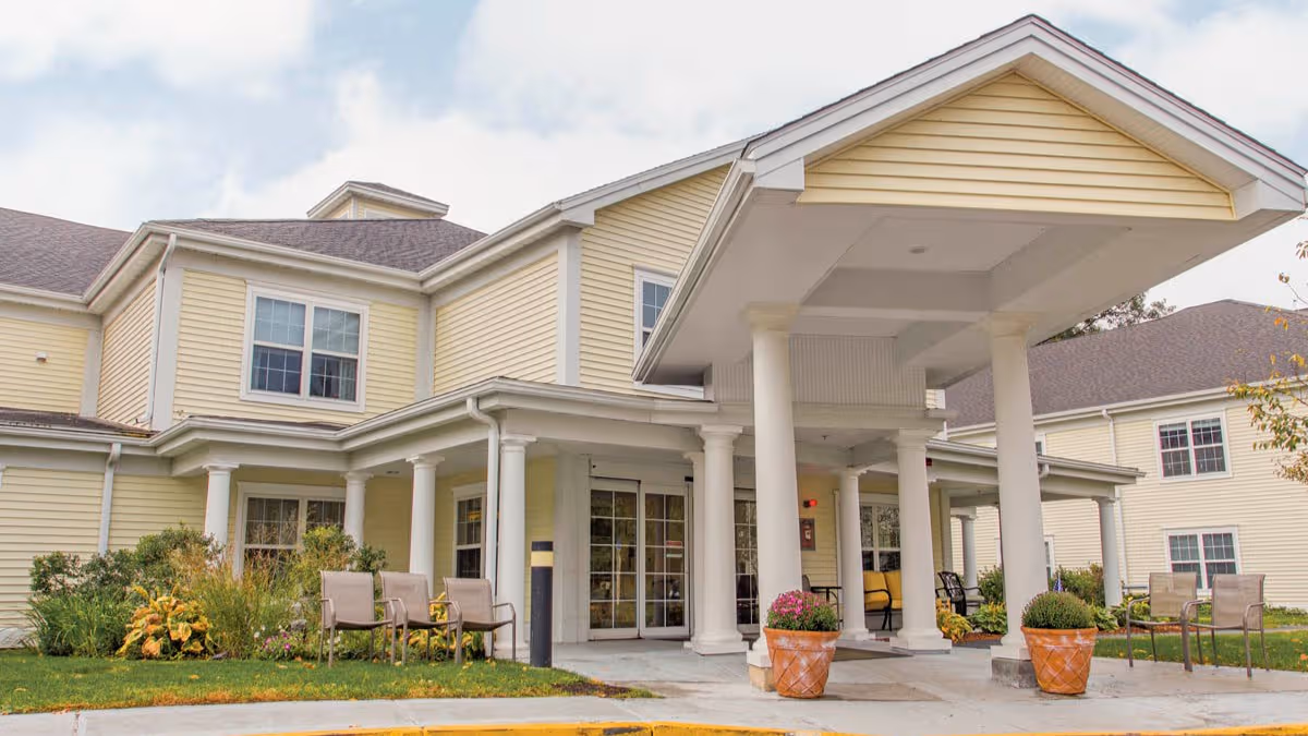 Yellow senior living building with a covered entrance and white columns, outdoor seating, and landscaping.