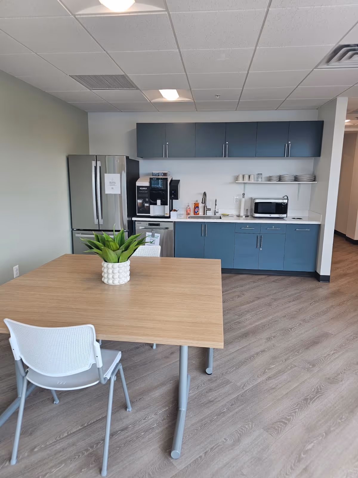 Modern kitchenette with blue cabinets, stainless refrigerator, coffee area, and wooden table.