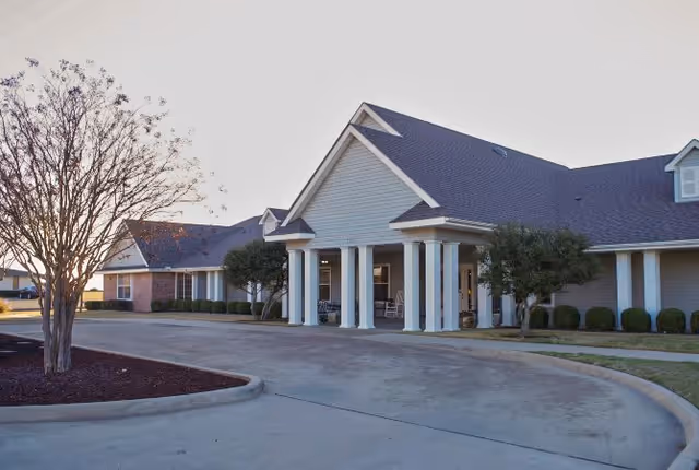 Exterior view of Wildflower Place facility showing a single-story building with a covered entrance supported by white columns, surrounded by trimmed bushes and trees, with a curved driveway in front.