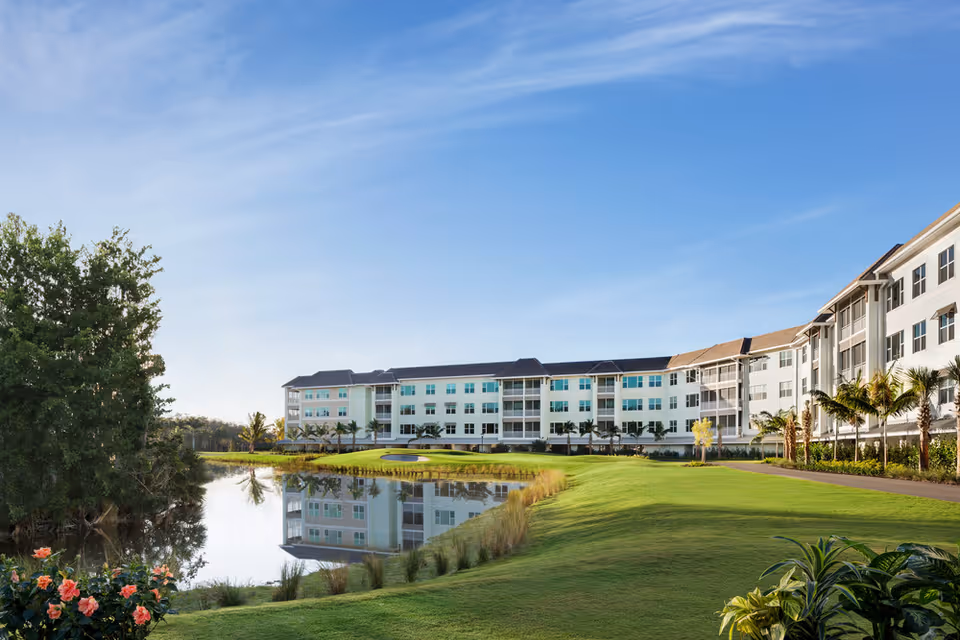 A large, modern senior living facility building with multiple floors, surrounded by well-maintained green lawns, palm trees, and a small pond reflecting the building. The sky is clear and blue.