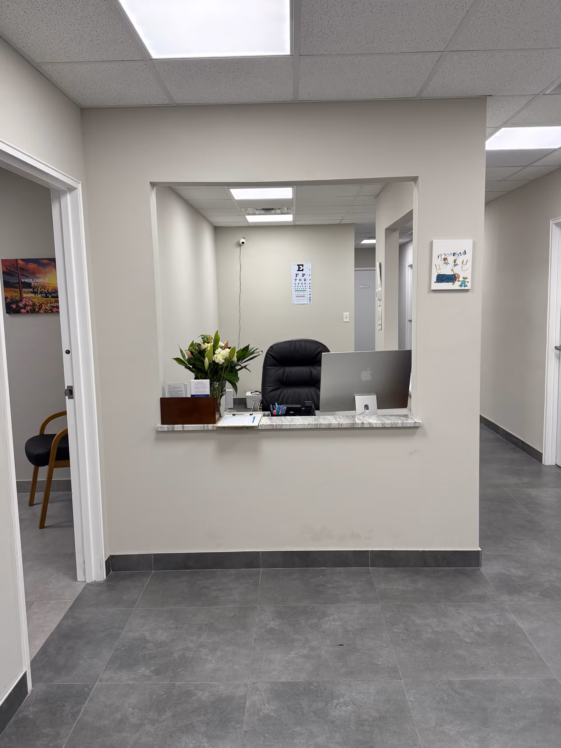 Interior reception desk with computer, flowers, and a black chair in a neutral hallway.