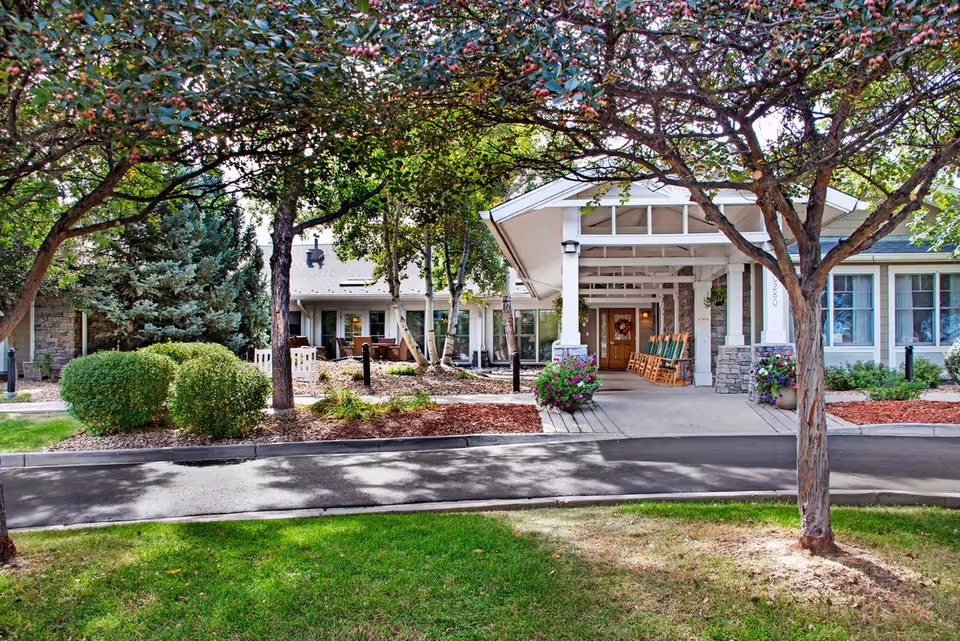 Front entrance of a senior living facility with a covered porte-cochere, rocking chairs on the porch, and landscaped trees and shrubs.
